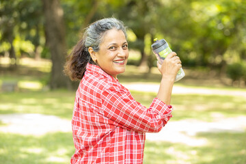 Happy fitness senior indian woman resting holding water bottle after work out exercising in park summer morning outdoor portrait.