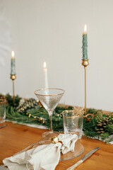 Christmas table setting. Linen napkin with bell on plate, vintage cutlery, glasses, fir branches with golden lights and pine cones on table. Holiday arrangement of table