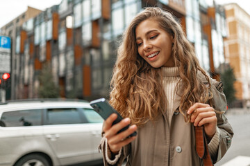 Smiling curly woman wearing warm coat walking down the street and using her phone