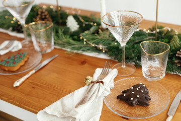 Christmas table setting. Christmas tree cookie on plate, linen napkin with bell, vintage cutlery, glasses, fir branches with golden lights and pine cones on table. Modern Holiday arrangement