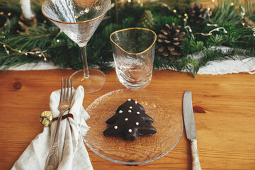 Fototapeta na wymiar Stylish Christmas table setting. Christmas tree cookie on plate, linen napkin with bell, vintage cutlery, wineglass, fir branches with golden lights, candles on table. Atmospheric Holiday brunch