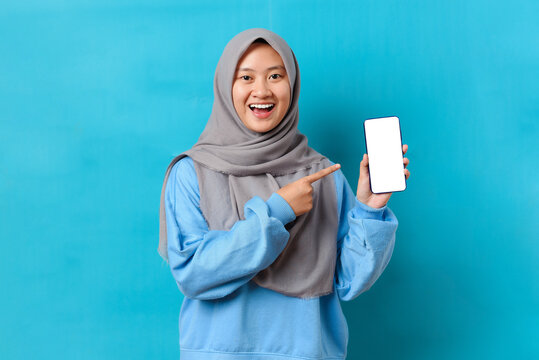 Happy beautiful young Indonesian woman with hijab showing blank white screen mobile phone and pointing finger at phone screen