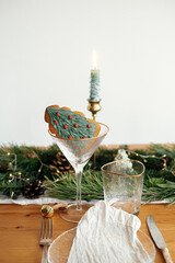 Stylish Christmas table setting. Christmas tree cookie in wineglass, plate with linen napkin, vintage cutlery, fir branches with golden lights, candles on table. Holiday brunch, creative table set