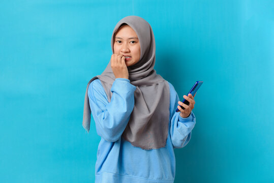 Photo of nervous scared Indonesian woman holding a smartphone with worried expression at camera, sees phobia, afraids of speaking, wears blue jumper