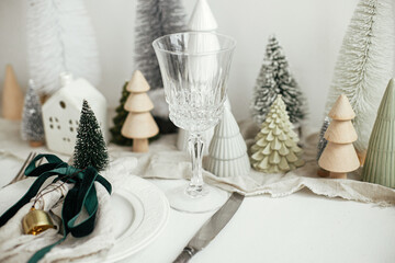 Stylish Christmas table setting. Festive napkin with ribbon and bell on plate, vintage cutlery, wineglass, modern christmas trees and houses on white rustic table. Holiday brunch