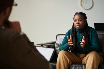 Young African American girl sitting on sofa and discussing her problems with psychologist at office