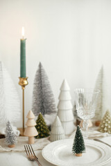 Modern Christmas table setting. Stylish little christmas tree on plate, vintage cutlery, glasses, festive fir and houses on white rustic table. Holiday arrangement of table