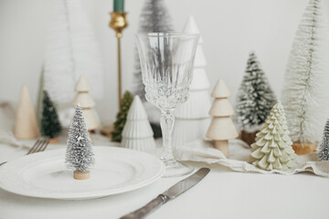Stylish Christmas table setting. Modern little christmas tree on plate, vintage cutlery, wineglass, modern christmas trees and houses on white rustic table. Holiday brunch, new year celebration