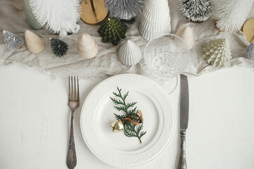 Fototapeta na wymiar Modern Christmas table setting. Cedar branch with bell on plate, vintage cutlery, glasses, festive little christmas trees and houses on white rustic table. Holiday arrangement of table, top view