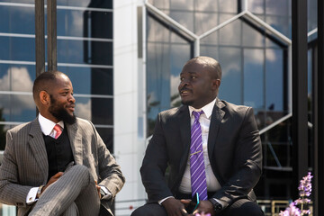 Two dark-skinned businessmen in suits communicate. Friends and business partners are sitting on a bench in the city against the backdrop of a glass building