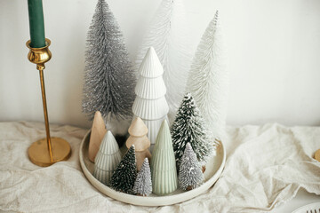 Stylish christmas little trees on plate, linen runner, golden candles on rustic table. Modern white artificial fir trees decoration in room. Holiday arrangement of table, christmas setting