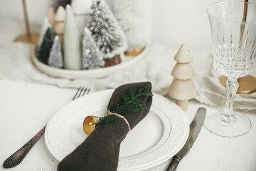 Christmas table setting. Stylish napkin with bell and fir on plate, vintage cutlery, glasses, festive little christmas trees on white rustic table. Holiday arrangement of table