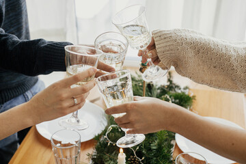 Christmas feast. Hands with wine glasses toasting and clinking on background of stylish table setting with fir branches with golden lights and candles. Friends celebrating with champagne