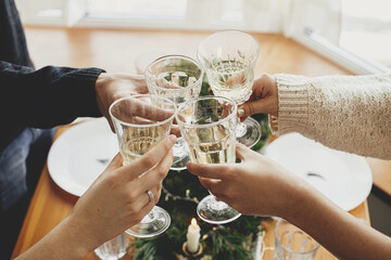 Christmas feast. Hands with wine glasses toasting and clinking on background of stylish table setting with fir branches with golden lights and candles. Friends celebrating with champagne