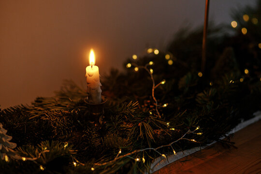Stylish christmas candle with fir branches and golden lights in evening dark room on table. Atmospheric christmas or new year's eve. Moody night image with golden lights and pine decor