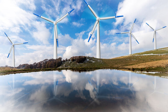 Group of white and blue wind turbines on a mountain landscape with a beautiful lake with reflections. Blue sky with clouds and sunbeams on background.