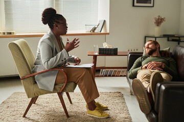 African American psychologist sitting on armchair and giving recommendation to patient while he lying on sofa during consultation at office - obrazy, fototapety, plakaty