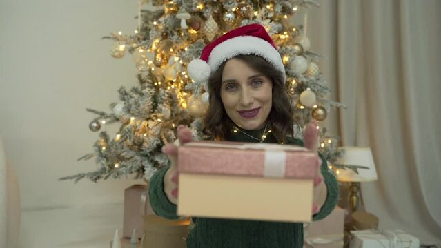 Woman wearing green sweater and Santa's cap presenting a gift near the tree