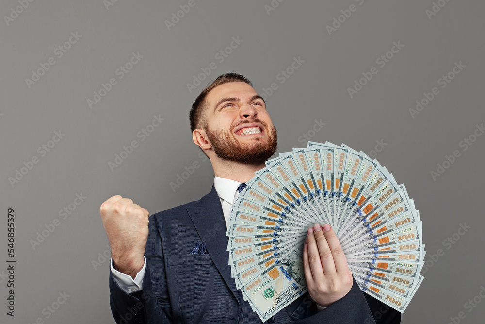 Wall mural excited businessman in suit with us dollars banknotes fan in hand against grey background. business 