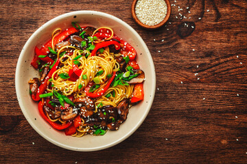 Vegan Stir fry noodles with vegetables, paprika, mushrooms, chives and sesame seeds in bowl. Wooden table background, top view, copy space