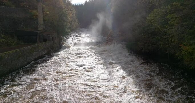 Slow motion drone footage slowly reversing down a fast flowing river and waterfall surrounded by old buildings. Autumnal trees overhang a gorge. Falls of Clyde, New Lanark, Scotland.