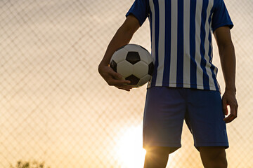 Athlete standing with ball on football field during sunrise, soccer ball in net on sky background,...