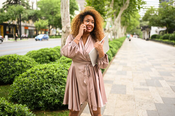 Attractive happy stylish plus size African American woman student freelancer afro hair with laptop computer outside. Young female office worker speaks phone in big urban city summer street. Diversity