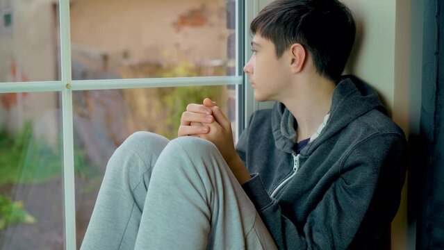 Scared sad boy refugee sits on windowsill and looks out window at curfew because russian armored invasion into Ukraine. Save lifes, no war, world peace, stop conflict, born love. Childhood in