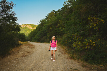 Excited happy senior woman backpacker tourist walking in summer forest road outdoors at sunset time. Old slim lady traveling with photo camera. Active retirement vacation concept. Warm filter