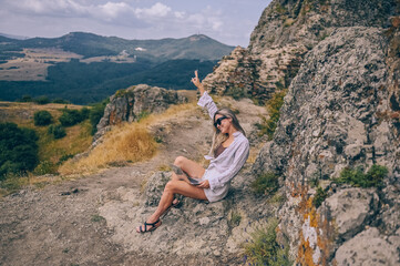 Naklejka premium Young woman freelancer traveler working online anywhere outdoors using laptop enjoying mountain peak view. Happy female downshifter in sunglasses holding computer in unusual workplace at summer