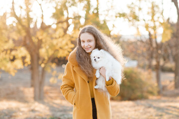 Cute smiling teenage girl 15-16 year old holding white fluffy spitz puppy dog together over autumn yellow nature background in park. Friendship.