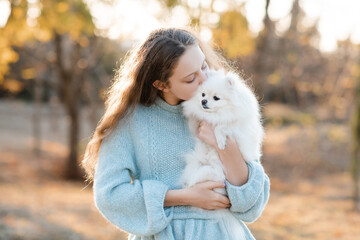 Cute smiling teenage girl 15-16 year old holding white fluffy spitz puppy dog together over autumn yellow nature background in park. Friendship.