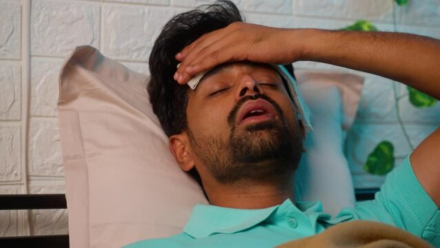young indian sick man placing wet cloth on forehead to reduce temperatureof fever while sleeping at night by - concept of healthcare, covid symptoms and illness.