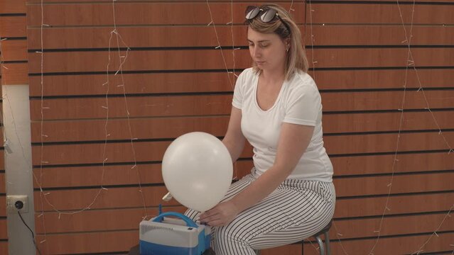 a girl sits on a chair and uses a balloon inflator to inflate white balloons