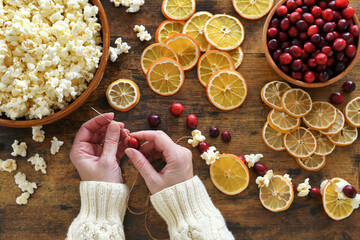 Female making homemade Christmas garland by stringing popcorn, cranberries, dried orange and lemon...