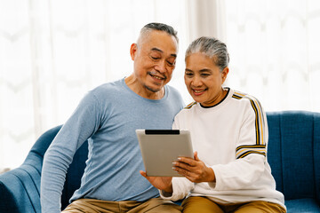 Smiling senior couple sitting on the couch looking at tablet computer, Asian retirees use...
