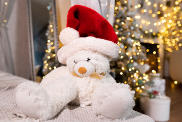 White teddy bear in a red santa claus hat sits on a bed against the backdrop of a luxuriously decorated Christmas tree. Evening on New Year's Eve, Christmas