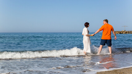 A happy man and woman are walking along sea smiling and holding hands.