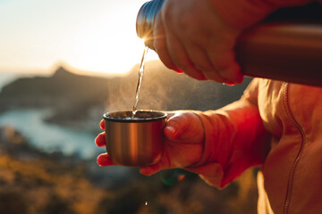 A human pours hot tea from a thermos into a mug on the mountain. Concept of autumn outdoor...