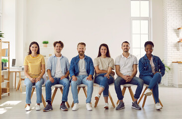 Portrait of smiling multiracial young people in casual clothes sitting in row on chairs. Cheerful diverse men and women in jeans, sneakers and t-shirts are sitting in row in room and looking at camera