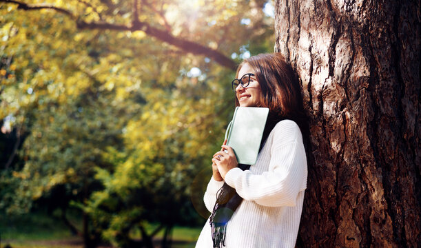 Woman in glasses holding book in her hands and smiling while walking outdoors in nature.