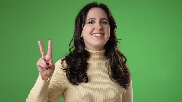 Portrait of happy young woman 20s smiling and giving peace or victory sign gesture with hands isolated on green screen background.