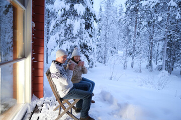 mature couple, man and woman are sitting on cozy wooden chairs on terrace of country house in winter forest and drinking hot tea, early evening, warm light from large window, enjoying winter nature