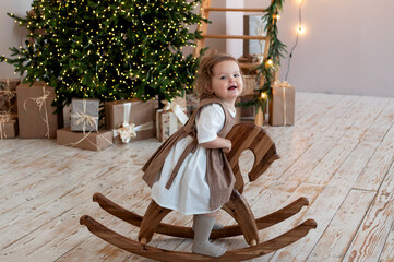 little girl swings on a wooden toy - a rocking chair at home against the backdrop of a Christmas...
