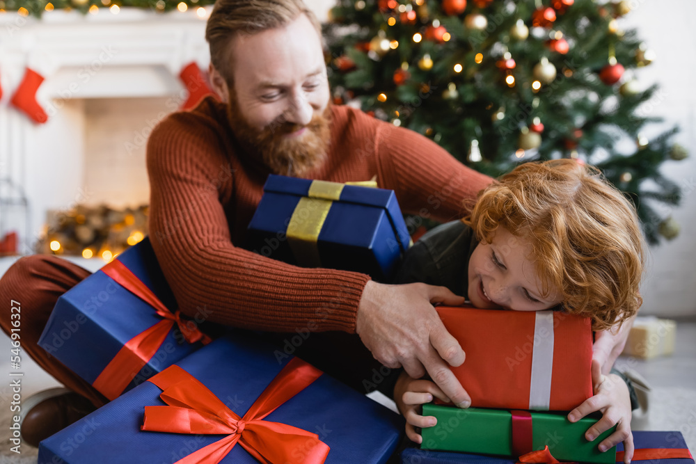 Wall mural cheerful bearded man and redhead child having fun with Christmas gift boxes at home