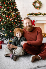 Obraz na płótnie Canvas cheerful father and child with red hair sitting on floor near Christmas tree and looking at camera