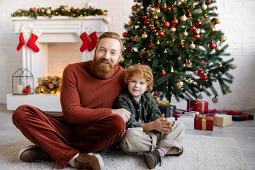 Obraz na płótnie Canvas redhead father and son sitting with crossed legs on floor in living room with Christmas tree and decorated fireplace