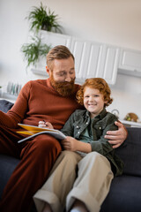 Obraz premium happy bearded man hugging cheerful redhead son sitting on couch with notebook