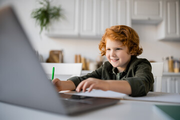 cheerful redhead boy doing homework near blurred laptop in kitchen