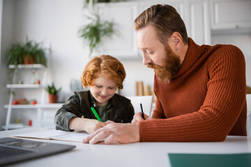 smiling kid writing in notebook near bearded dad and laptop on blurred foreground